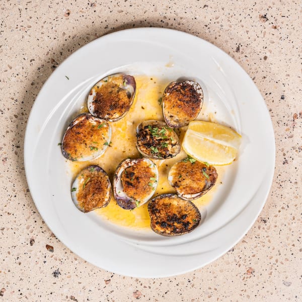 White plate holds baked clams with golden breadcrumb topping, lemon, and butter sauce.