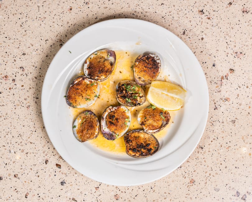 White plate holds baked clams with golden breadcrumb topping, lemon, and butter sauce.