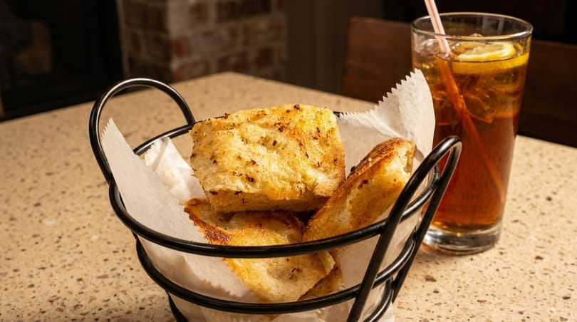 Basket of golden-brown focaccia bread with herbs and a glass of iced tea with lemon.